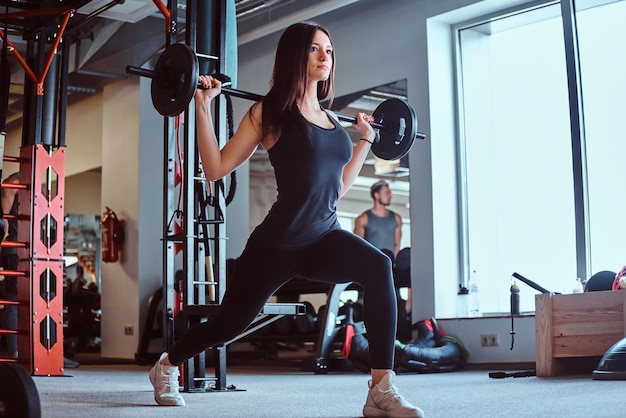 Bella bruna femmina in abiti sportivi facendo affondo con bilanciere nel fitness club o in palestra.