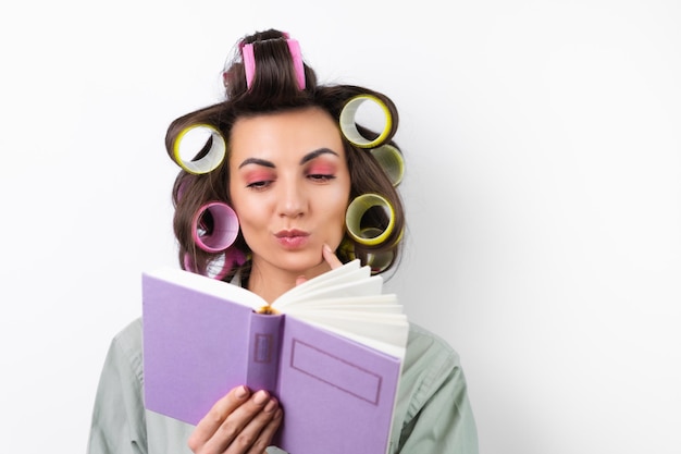 Bella casalinga Giovane donna allegra con bigodini trucco luminoso con un libro in mano su uno sfondo bianco Pensando a una ricetta per la cena Alla ricerca di idee alimentari