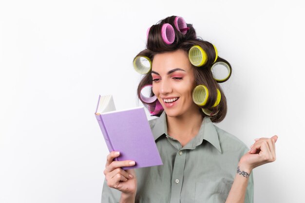 Bella casalinga Giovane donna allegra con bigodini trucco luminoso con un libro in mano su uno sfondo bianco Pensando a una ricetta per la cena Alla ricerca di idee alimentari