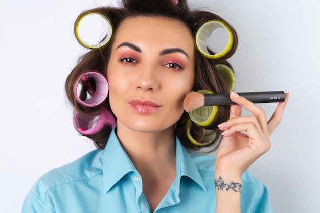 Bella casalinga giovane donna allegra con bigodini trucco rosa brillante e una spazzola da cotouring si sta preparando per una cena con appuntamento notturno su sfondo bianco