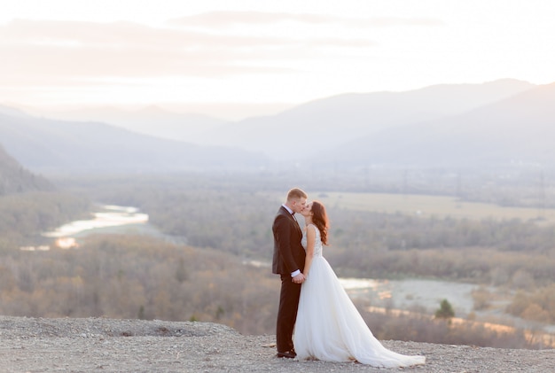Bella coppia di sposi bacia sulla collina in vista di un paesaggio pittoresco nel crepuscolo