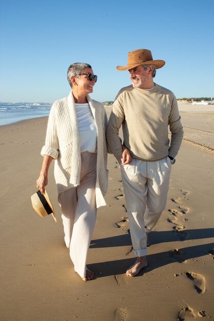 Bella coppia senior che passeggia a piedi nudi lungo la spiaggia lasciando dietro di sé dei passi sulla sabbia bagnata. Belle coppie che si godono le vacanze in riva al mare, camminando tenendosi per mano. Amore, concetto di vacanza