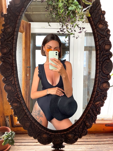 Bella donna a casa scatta foto selfie allo specchio sul cellulare per storie e post sui social media, indossando un costume da bagno estivo nero