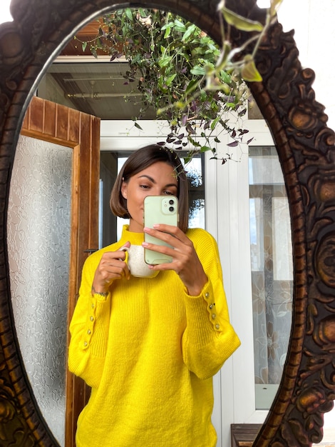 Bella donna a casa scatta foto selfie allo specchio sul telefono cellulare per storie e post sui social media, indossando un maglione giallo caldo e accogliente