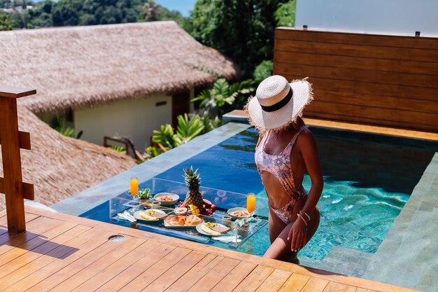 Bella donna abbronzata caucasica in bikini e cappello di paglia con colazione galleggiante in incredibile villa di lusso in stile bali al giorno pieno di sole in piscina, sfondo tropicale.