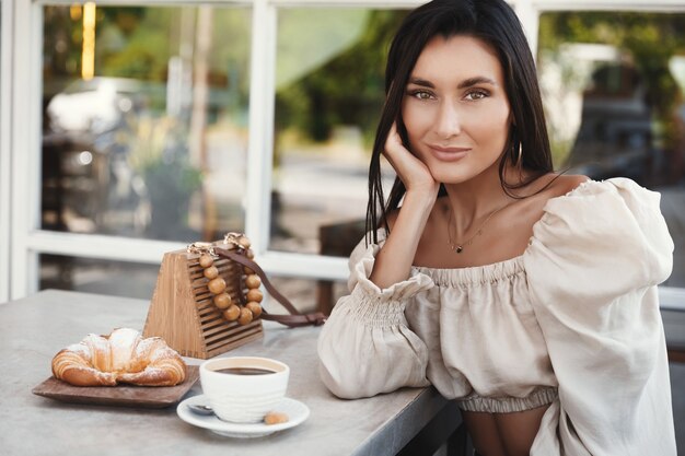 Bella donna abbronzata in camicetta elegante, bere caffè in un bar sorridendo alla telecamera.