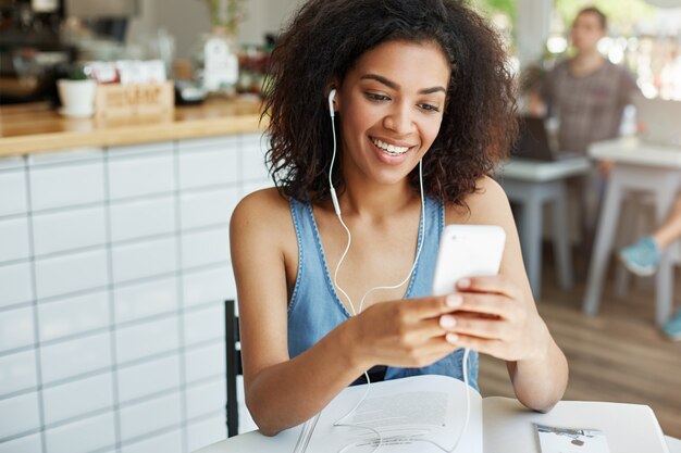 Bella donna africana in cuffie che sorride guardando lo schermo del telefono che si siede di riposo nella caffetteria