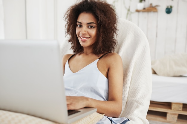 Bella donna africana nella seduta sorridente degli indumenti da notte nella sedia con il computer portatile a casa.