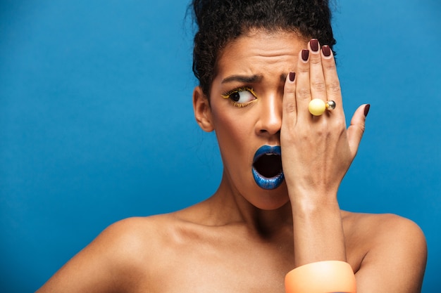 bella donna afro-americana con il trucco colorato che esprime eccitazione o sorpresa che copre un occhio con la mano, isolato su muro blu