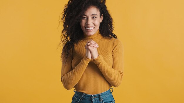 Bella donna afro eccitata con capelli scuri e soffici vestita con abbigliamento casual che sembra gioiosa sulla fotocamera su sfondo arancione