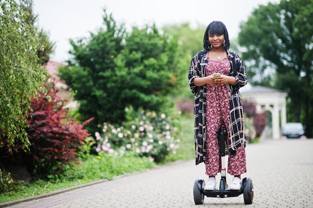 Bella donna afroamericana che usa segway o hoverboard Ragazza nera su scooter elettrico autobilanciante a doppia ruota con telefono cellulare a portata di mano