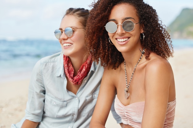 Bella donna afroamericana con pelle scura e capelli ricci, ha un sorriso positivo, indossa occhiali da sole, si siede vicino alla sua ragazza che ama il paesaggio marino, gode di un'atmosfera tranquilla in spiaggia. Concetto di amicizia