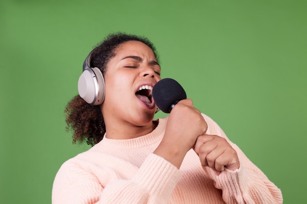 Bella donna afroamericana sulla parete verde con cuffie wireless e microfono cantando canzoni in karaoke