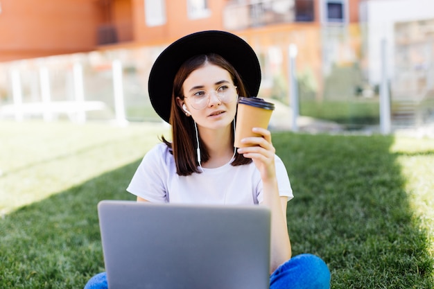 Bella donna alla moda che si siede sull'erba verde con il computer portatile e il caffè nella mano. Concetto di stile di vita