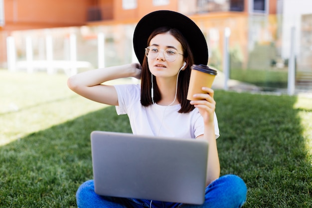 Bella donna alla moda che si siede sull'erba verde con il computer portatile e il caffè nella mano. Concetto di stile di vita