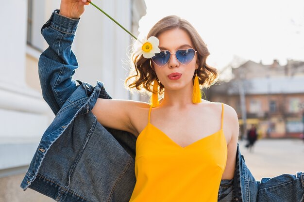 Bella donna alla moda hipster che si diverte, moda di strada, fiore in mano, vestito giallo, giacca di jeans, stile boho, tendenza moda primavera estate, occhiali da sole, sorridente, soleggiato, civettuolo