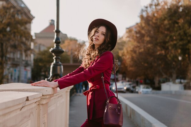 Bella donna alla moda in vestito viola che cammina nella via della città, tendenza di moda primavera estate autunno stagione indossando il cappello, tenendo la borsa