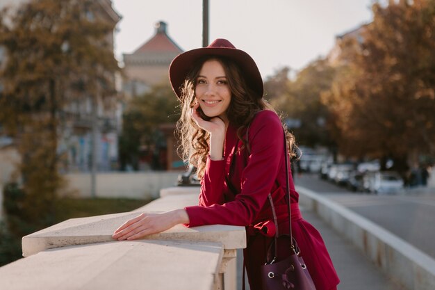 Bella donna alla moda in vestito viola che cammina nella via della città, tendenza di moda primavera estate autunno stagione indossando il cappello, tenendo la borsa