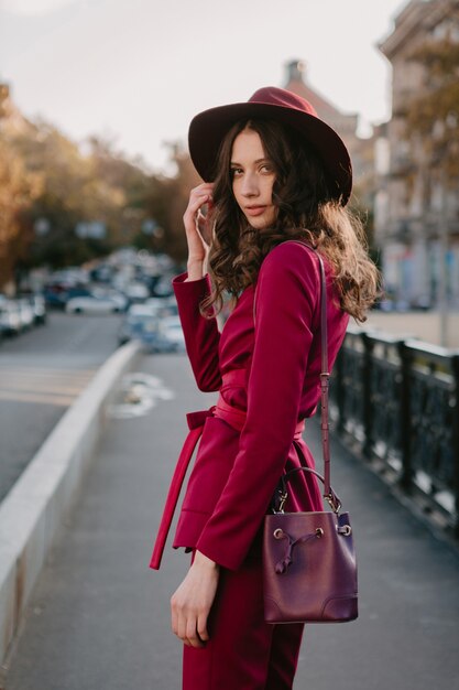 Bella donna alla moda in vestito viola che cammina nella via della città, tendenza di moda primavera estate autunno stagione indossando il cappello, tenendo la borsa