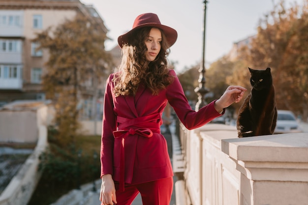 Bella donna alla moda in vestito viola in via della città, tendenza di moda primavera estate autunno stagione indossa il cappello, guardando un gatto