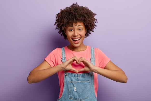 Bella donna allegra con la pelle scura, taglio di capelli afro mostra un gesto a forma di cuore, indossa una maglietta casual e una tuta di jeans