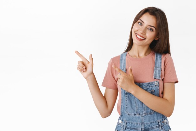 Bella donna allegra e seducente in salopette tshirt inclina la testa sciocca e sorridente mentre promuove il prodotto dell'azienda rivolto verso l'angolo in alto a sinistra dà consigli per dare un'occhiata alla pubblicità interessante