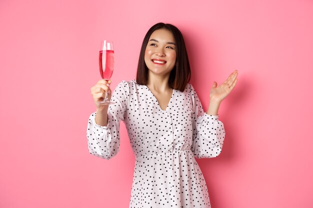 Bella donna asiatica, alzando un bicchiere di champagne e sorridendo felice, bevendo vino e festeggiando, in piedi su sfondo rosa.