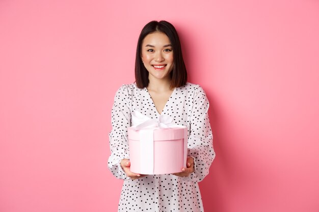 Bella donna asiatica che augura buone feste dandoti un regalo in una scatola carina in piedi su sfondo rosa...