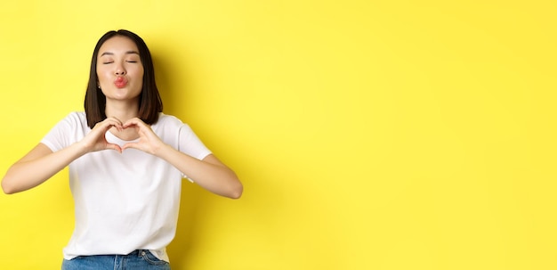 Bella donna asiatica che mostra ti amo gesto del cuore sorridendo alla telecamera in piedi contro il bac giallo