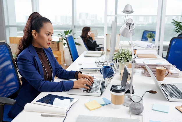 Bella donna asiatica che si siede allo scrittorio in ufficio occupato e che lavora al computer portatile