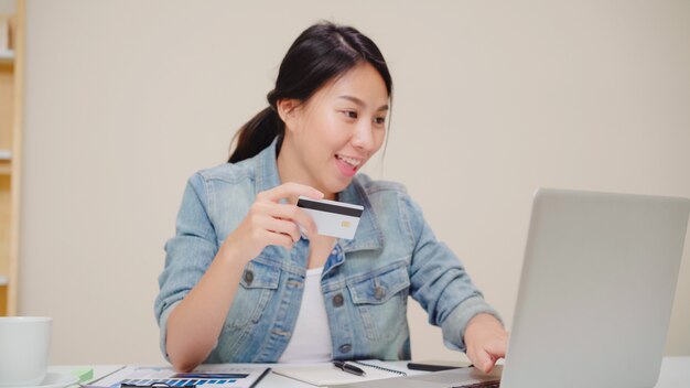 Bella donna asiatica che utilizza computer portatile che compra acquisto online dalla carta di credito mentre usura seduta casuale sullo scrittorio in salone a casa.
