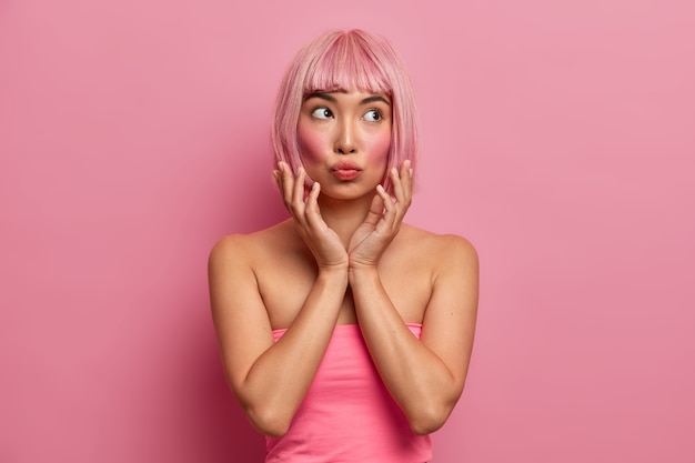 Bella donna asiatica con guance rosse, tiene le labbra piegate, mani vicino al viso, ha un'espressione pensierosa, capelli rosa, vestita in alto