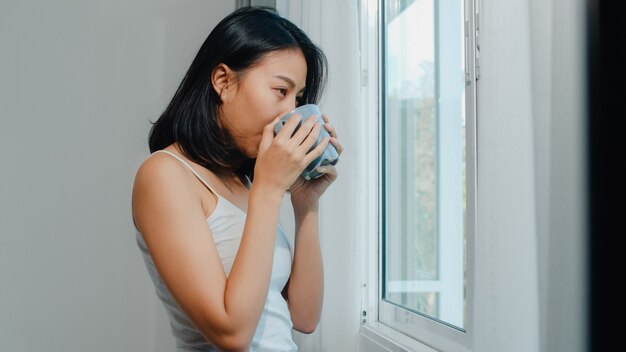 Bella donna asiatica felice che sorride e che beve una tazza di caffè o un tè vicino alla finestra in camera da letto. Le tende aperte della giovane ragazza latina e si rilassano nella mattina. Stile di vita signora a casa.