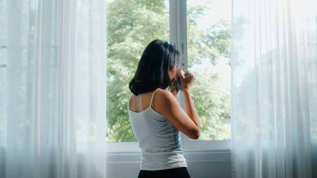 Bella donna asiatica felice che sorride e che beve una tazza di caffè o un tè vicino alla finestra in camera da letto. Le tende aperte della giovane ragazza latina e si rilassano nella mattina. Stile di vita signora a casa.