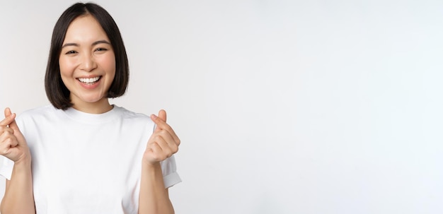 Bella donna asiatica sorridente che mostra i cuori del dito gesto che indossa la maglietta in piedi su sfondo bianco