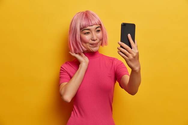 Bella donna asiatica utilizza gadget per videochiamata, corregge i capelli rosa, guarda la fotocamera dello smartphone, fa selfie