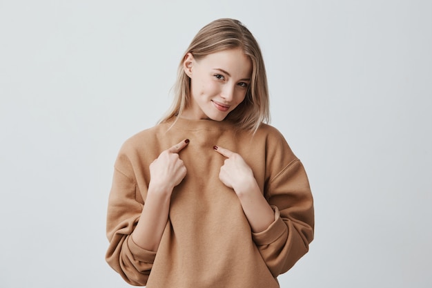 bella donna bionda attraente che sorride, indicando se stessa con le dita, vestita in maglione a maniche lunghe beige, che esprime emozioni e sentimenti positivi.