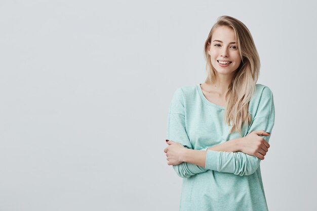 bella donna bionda con denti perfetti e pelle sana e pulita che riposa in casa, sorridendo felicemente dopo aver ricevuto buone notizie positive. Bella giovane donna in piedi con le braccia conserte