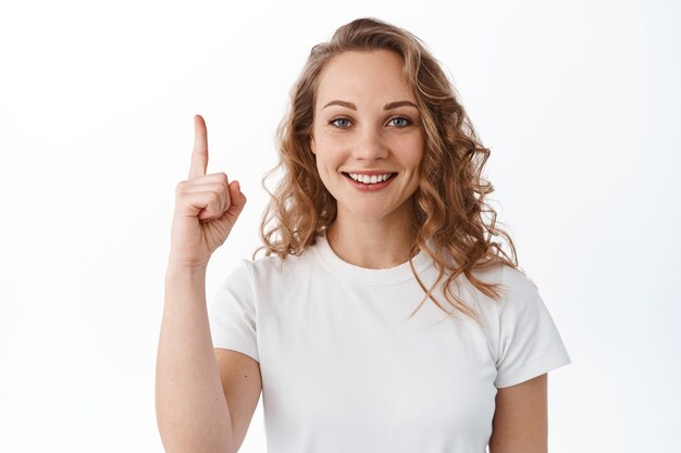 Bella donna bionda con l'acconciatura riccia, puntando il dito verso l'alto e sorridente, mostrando pubblicità, in piedi sul muro bianco