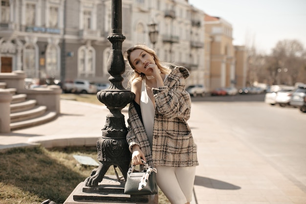 Bella donna bionda in abito di seta e cappotto di tweed a scacchi di grandi dimensioni si appoggia al lampione tiene la borsetta e posa all'esterno