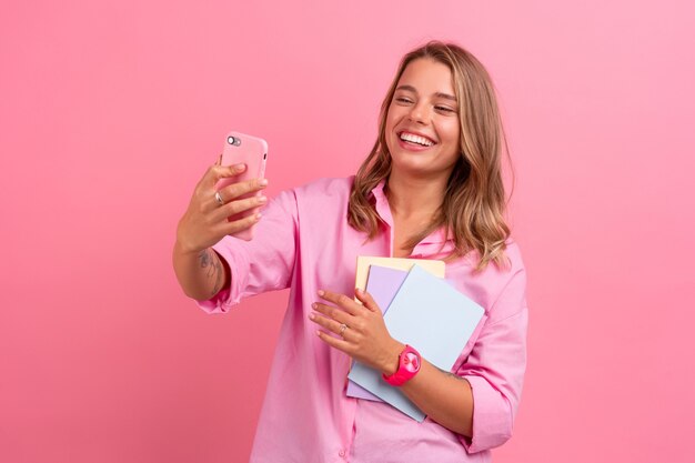 Bella donna bionda in camicia rosa sorridente che tiene in mano i taccuini e usa lo smartphone in posa sul rosa isolato