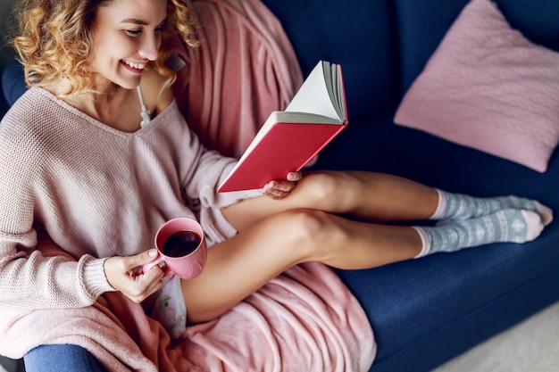 Bella donna bionda sorridente che si distende sullo strato a casa. Maglione lavorato a maglia rosa, calzini caldi. Libro di lettura e tazza di caffè della holding.