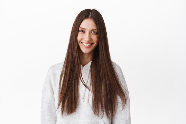 Bella donna bruna con lunghi capelli lucidi sani sorridente e guardando felice pubblicità di cosmetici per la cura dei capelli e di bellezza in piedi in felpa con cappuccio su sfondo bianco