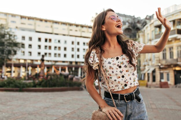 Bella donna bruna riccia in denim pantaloni elegante camicetta floreale e occhiali da sole rosa sorride sinceramente e saluta con le onde la mano all'aperto