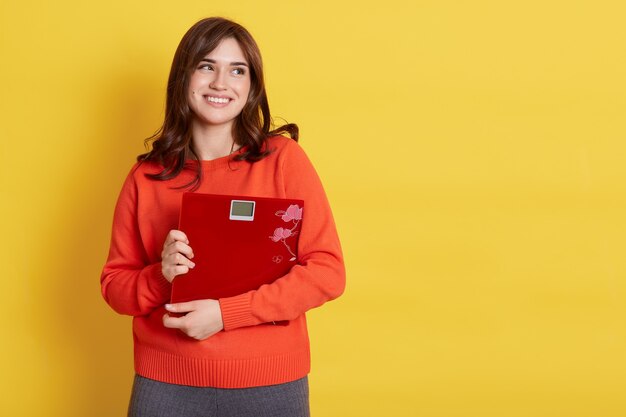 Bella donna bruna sorridente in maglione casual arancione che abbraccia bilance da pavimento e distoglie lo sguardo con espressione sognante, isolata sopra la parete gialla.