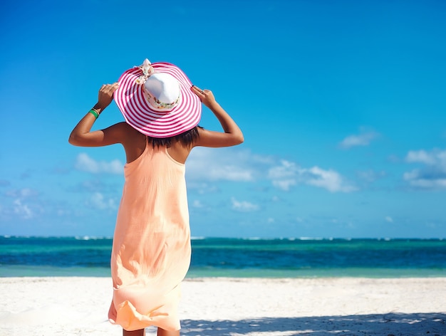 Bella donna calda in cappellino da sole variopinto e vestito che camminano vicino all'oceano della spiaggia il giorno di estate caldo sulla sabbia bianca