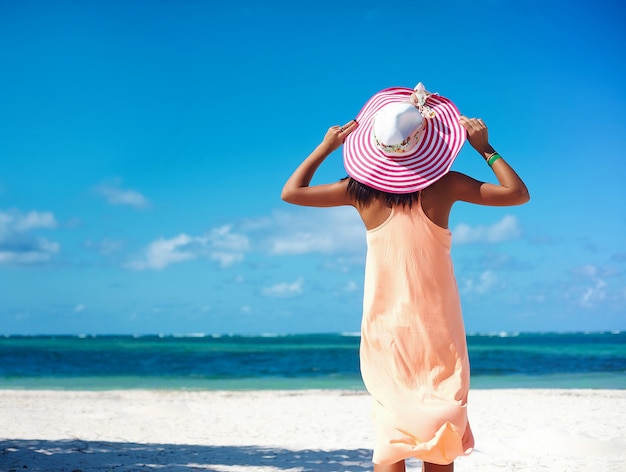 Bella donna calda in cappellino da sole variopinto e vestito che camminano vicino all'oceano della spiaggia il giorno di estate caldo sulla sabbia bianca