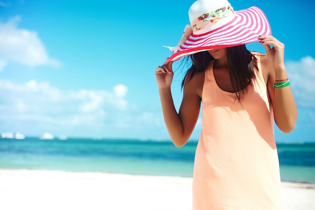 Bella donna calda in cappellino da sole variopinto e vestito che camminano vicino all'oceano della spiaggia il giorno di estate caldo sulla sabbia bianca