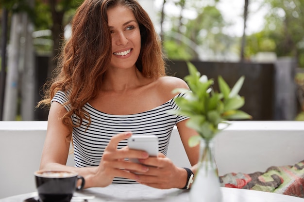 Bella donna castana con espressione felice e telefono nella caffetteria con terrazza all'aperto