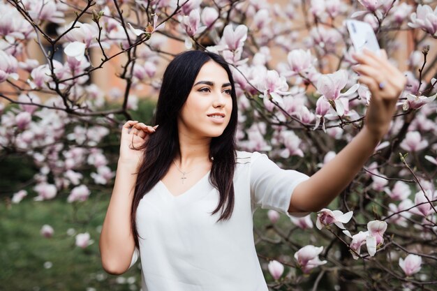 Bella donna caucasica alla moda che fa selfie nel giardino della magnolia del fiore. Vista dal basso